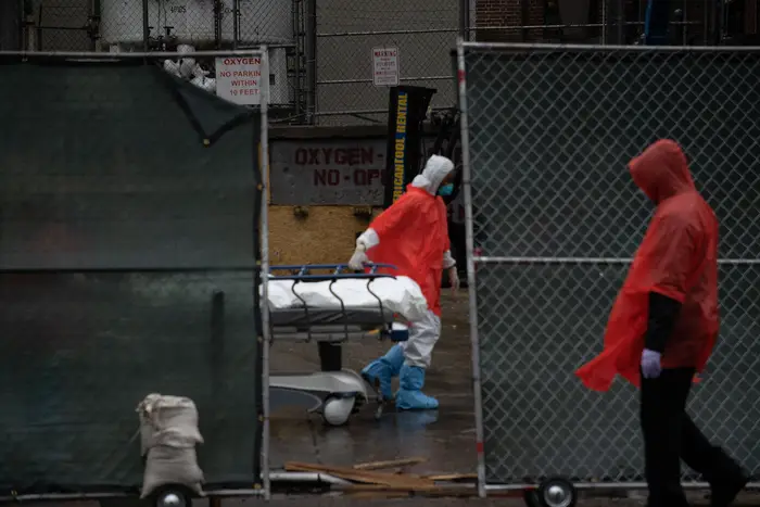 A worker behind a partially opened gates is seen in personal protective equipment pulling a stretcher with a body. Another worker is outside the gate.